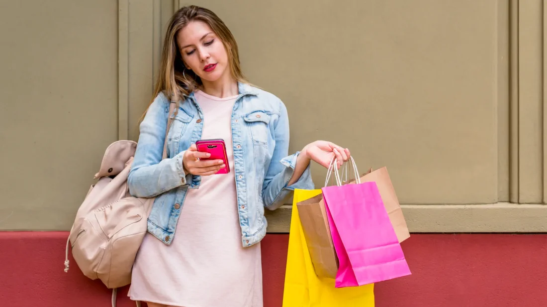 Woman with bright shein shopping bags using smartphone at building wall